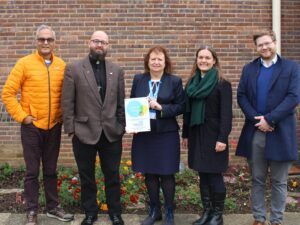 Five smiling people are standing outdoors, in front of a brick wall. The woman in the middle, who wears an NHS badge, is holding a certificate which says "We are a Living Wage Employer."