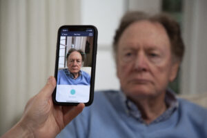 An older man is sitting in a room with a window and curtains behind him. Someone is holding a mobile phone in front of his face, which appears on the screen surrounded by a square frame. The phone is assessing his expression through facial recognition technology.