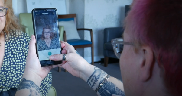 A woman is pointing a mobile phone at another woman's face, with the camera on. They are sitting in a lounge in a nursing home, and there are some armchairs in the background.