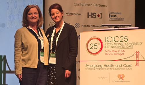 Two women dressed in business attire stand next to a podium which shows the name and dates of an academic conference in Lisbon, May 2025.
