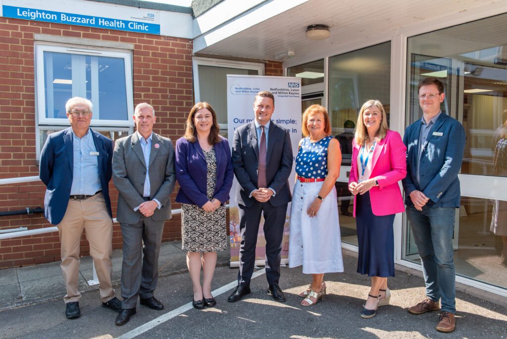 Seven people in business attire are standing in front of a brick building which bears the sign 'Leighton Buzzard Health Clinic'. They are smiling and look pleased.