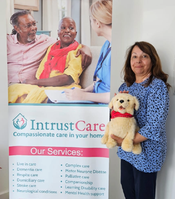 A woman stands in front of a banner reading 'Intrust Care', holding a medium-sized robotic dog. Both the woman and the dog are smiling.
