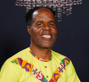 A man of Black heritage smiles at the camera.  He is wearing a brightly coloured shirt, which is mainly yellow.