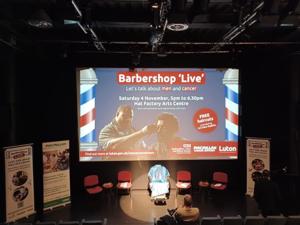 A spotlight on an empty chair on a stage, which symbolises the men living with prostate cancer who have not yet been diagnosed. Behind, there is a slideshow which reads 'Barbershop Live'.