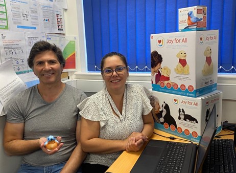 A man and a woman are smiling while sitting at a desk. He is holding a small robotic pet bird in his hand. Behind her, there is a stack of three boxes containing more robotic pets - a cat and a dog, as well as that for the bird.