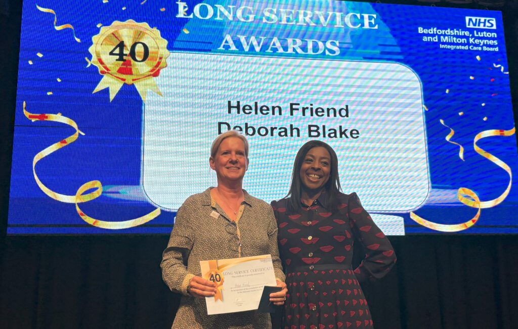 Two women are standing in front of a screen which says "Long Service Awards" with two names underneath. They are smiling. One of them is holding a certificate in her hands.