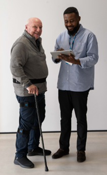 A man with an NHS lanyard writes something on a pad while he talks to an older man, who is using a walking stick and wearing some electronic sensors. They are fixed to his trousers above and on his knees, and on his ankles.