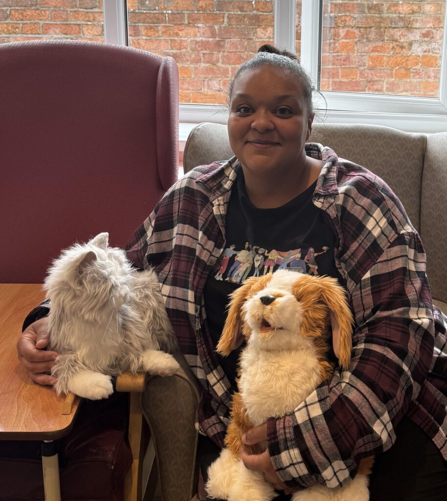A woman smiles at the camera as she holds a robotic cat and dog in her lap.