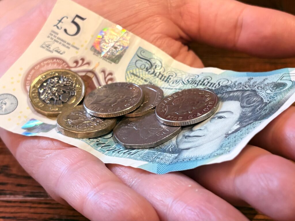 A hand holding a £5 note and some coins.