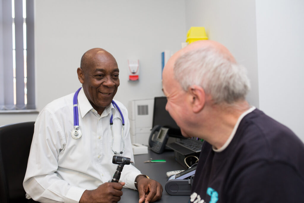 A man with a stethoscope around his neck and an otoscope in his hand is talking to another man. They are sitting in an office together.