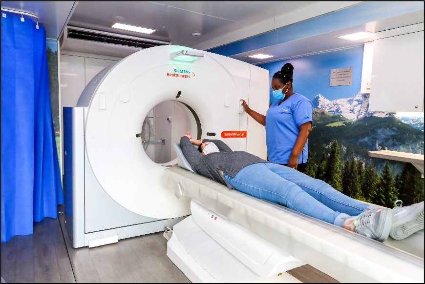 A person is lying on a surface with their arms above their head, waiting to enter a medical scanning machine. A nurse is standing alongside and seems to be explaining the process.